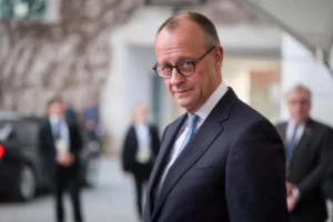 Фрідріх Мерц у костюмі та окулярах стоїть біля будівлі на тлі людей