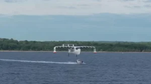 Білий екраноплан Regent Viceroy летить низько над водою біля берега, під ним катер