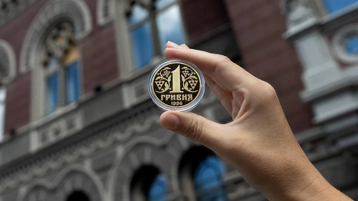 Рука тримає золотисту монету 1 гривня 1996 року на тлі історичної будівлі